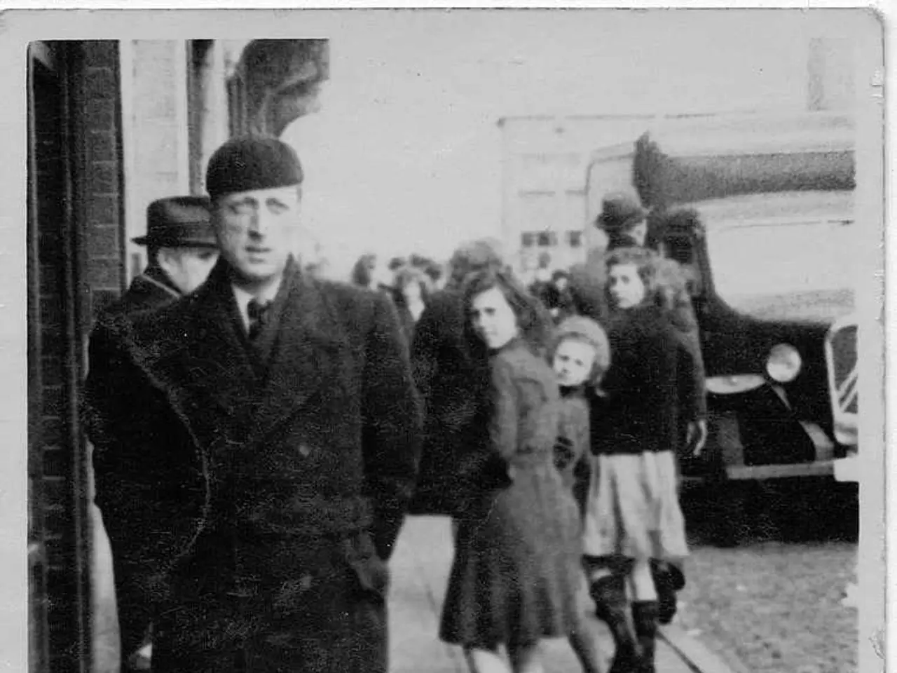 Black and white picture. Here we can see people and vehicle. Few people wore hats. Far there is a...