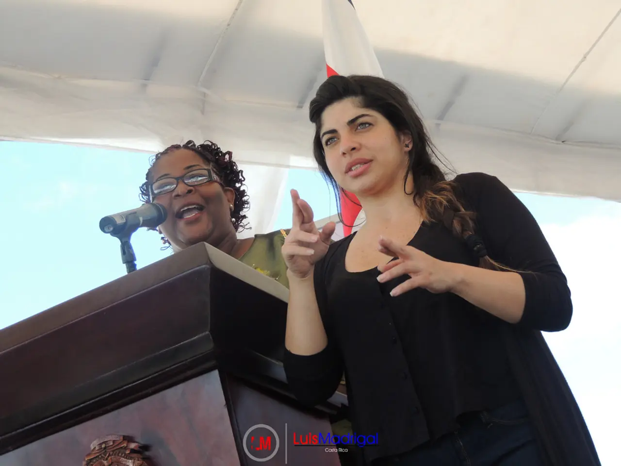 In this image we can see one white tent, one flag behind the human, two women standing near the...