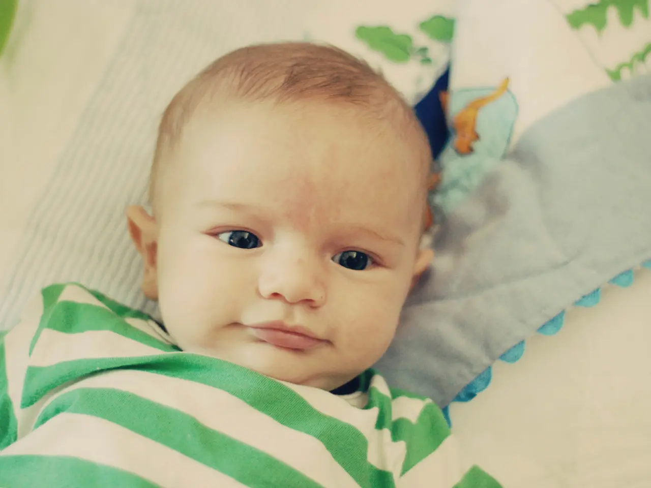 In the image there is a baby in white and green striped t-shirt laying on bed.
