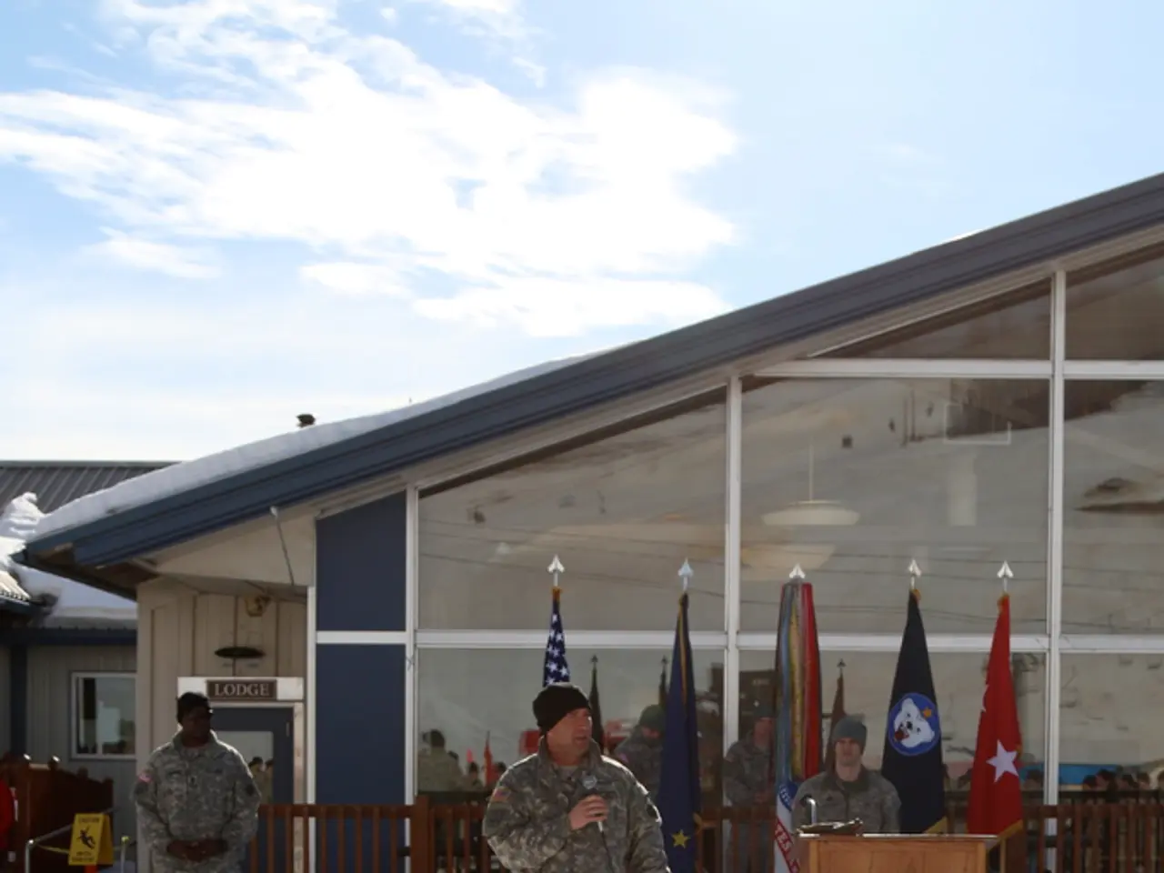 In this image we can see few people, flags, fence, boards, glass, and sheds. There is a podium. In...