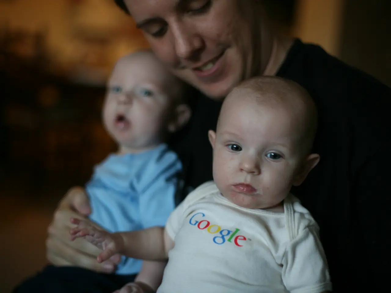 There is a person holding two babies. On the dress of the front baby something is written. In the...