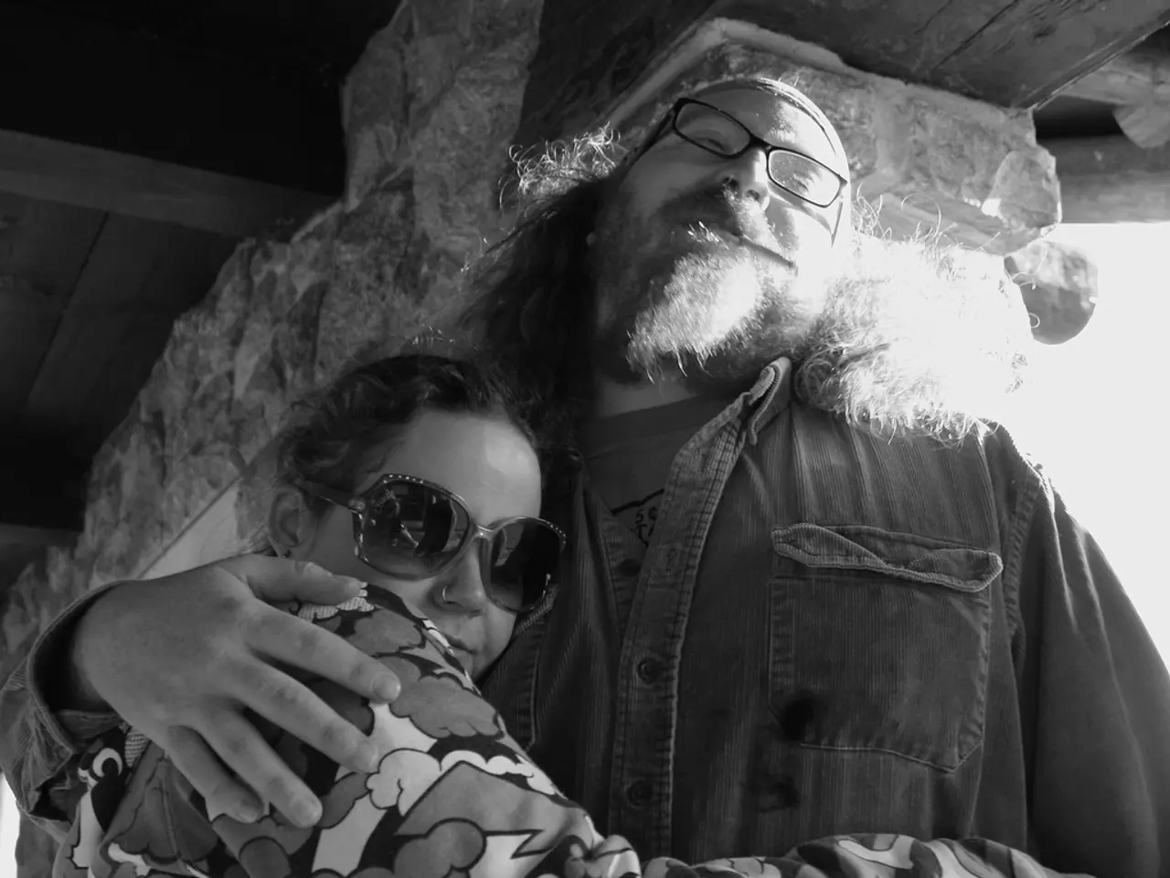 In the image I can see two people wearing spectacles and hugging each other and behind there is a...