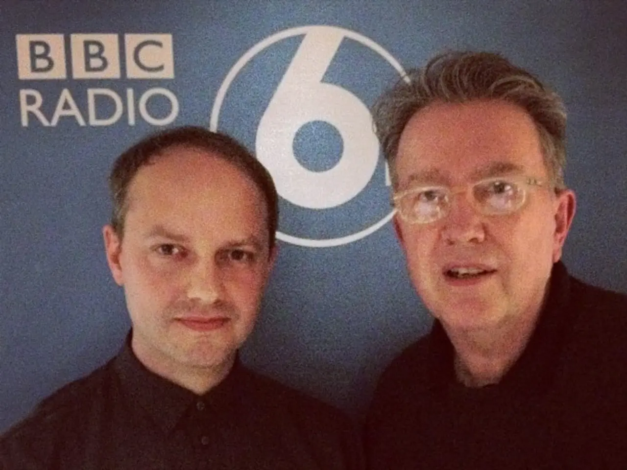 In this image I see 2 men who are wearing black dress and in the background I see the blue wall on...