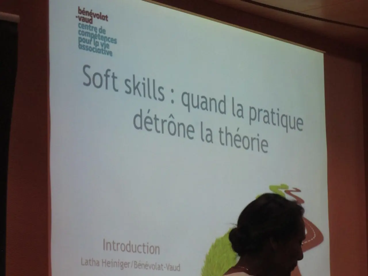 The woman in front of the picture is wearing a white T-shirt. She is holding a microphone in one of...