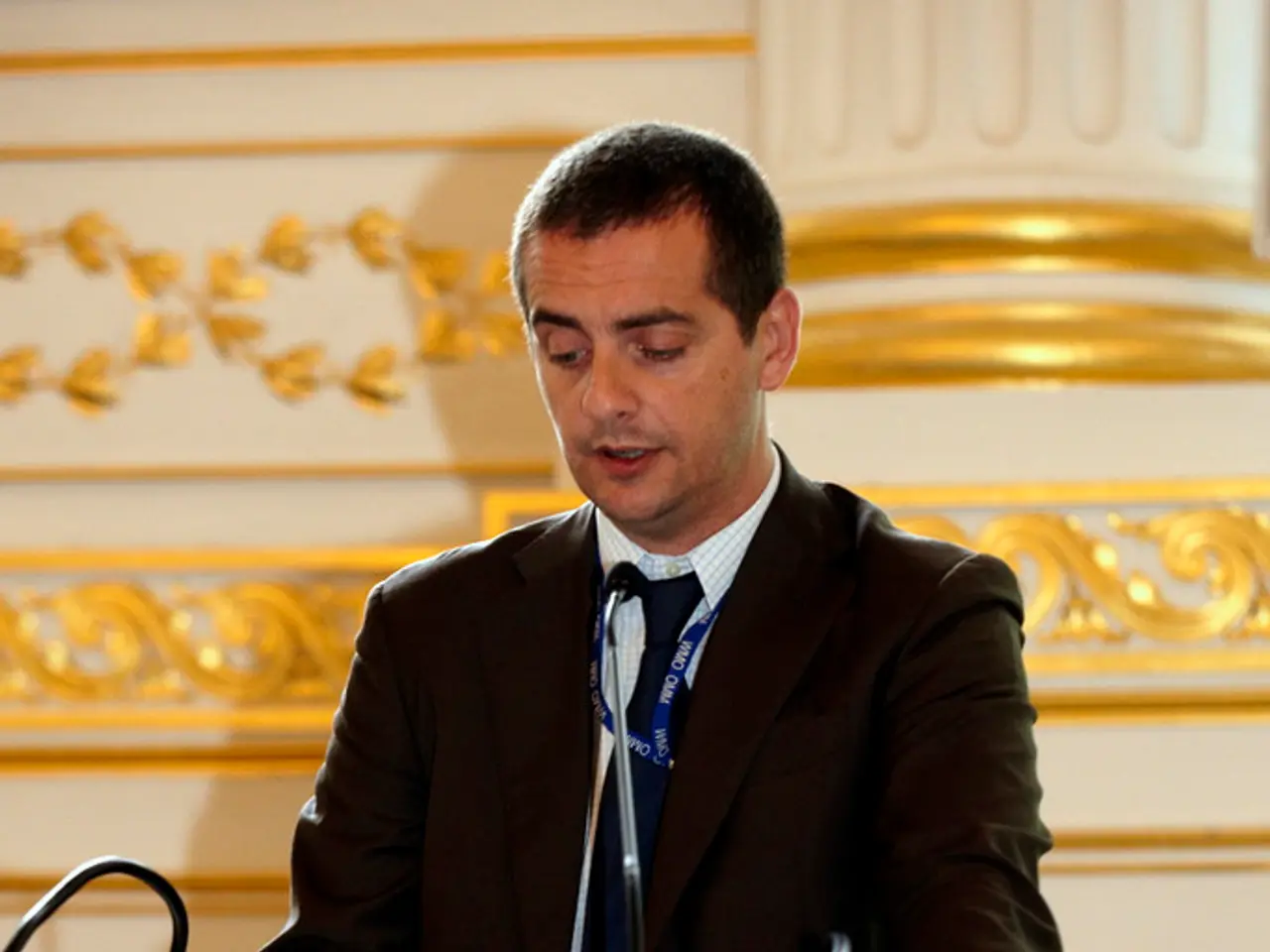 In this picture there is a man who is wearing suit. He is standing near to the speech desk. On the...