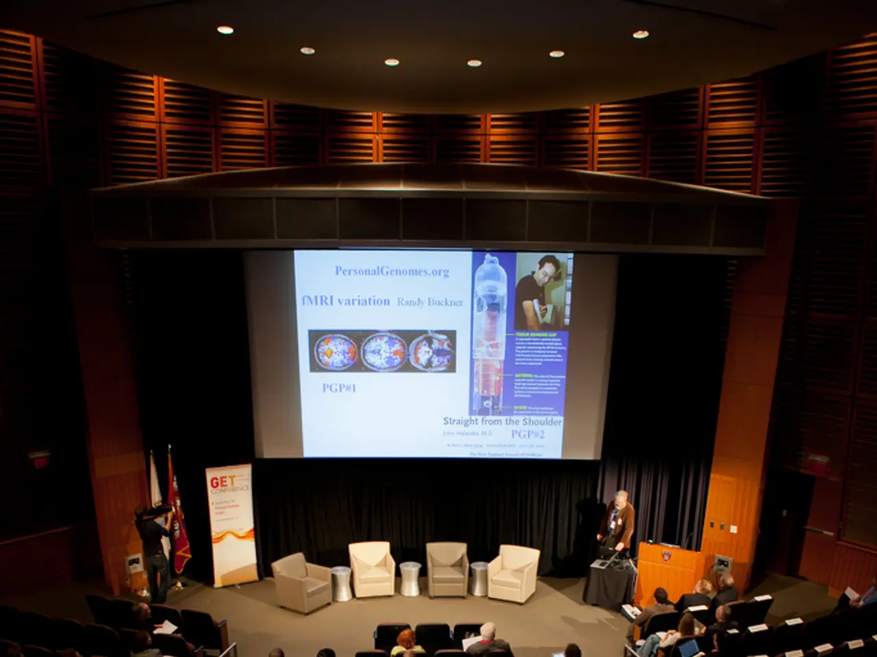 In this image we can see an auditorium. There are many people sitting on the chairs and holding...