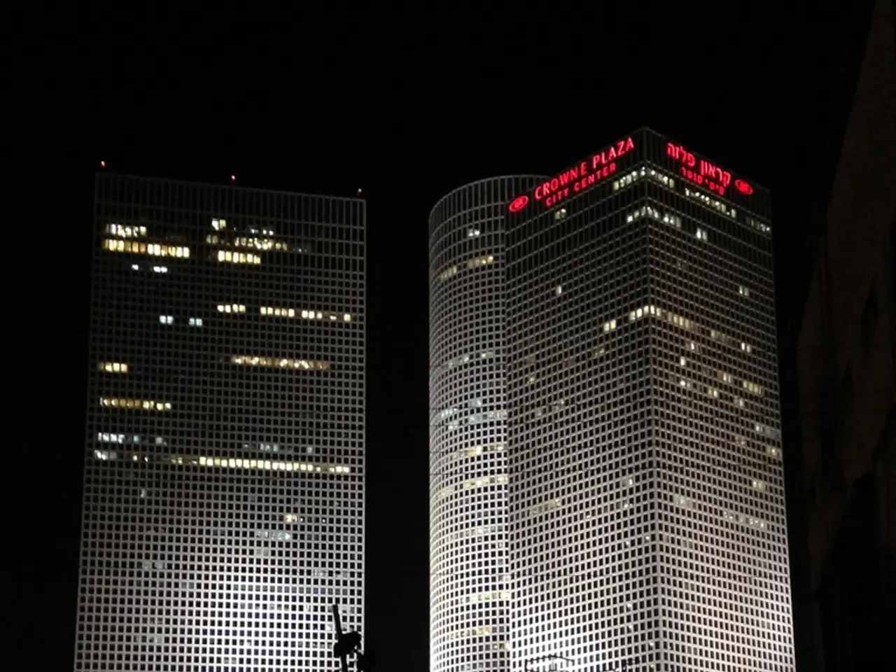 In this image there are few trees. Behind there are few buildings.