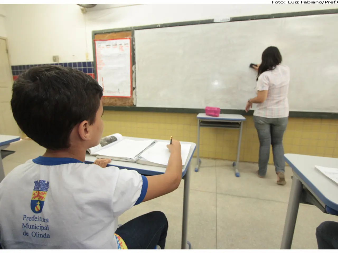 This picture shows a classroom. We see a boy seated and writing in the book on the table and we see...
