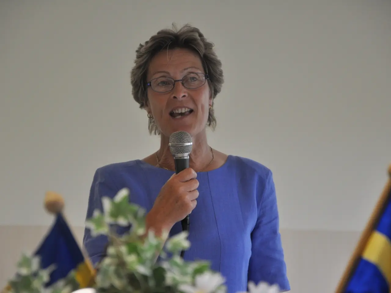 This picture shows a woman standing and speaking with the help of a microphone and we see two Flags...