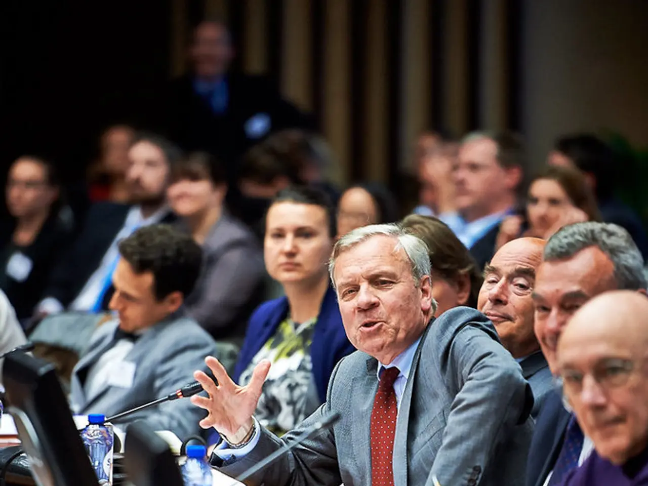 People are seated. There is a table on which there are screens, bottles, glasses, papers and...