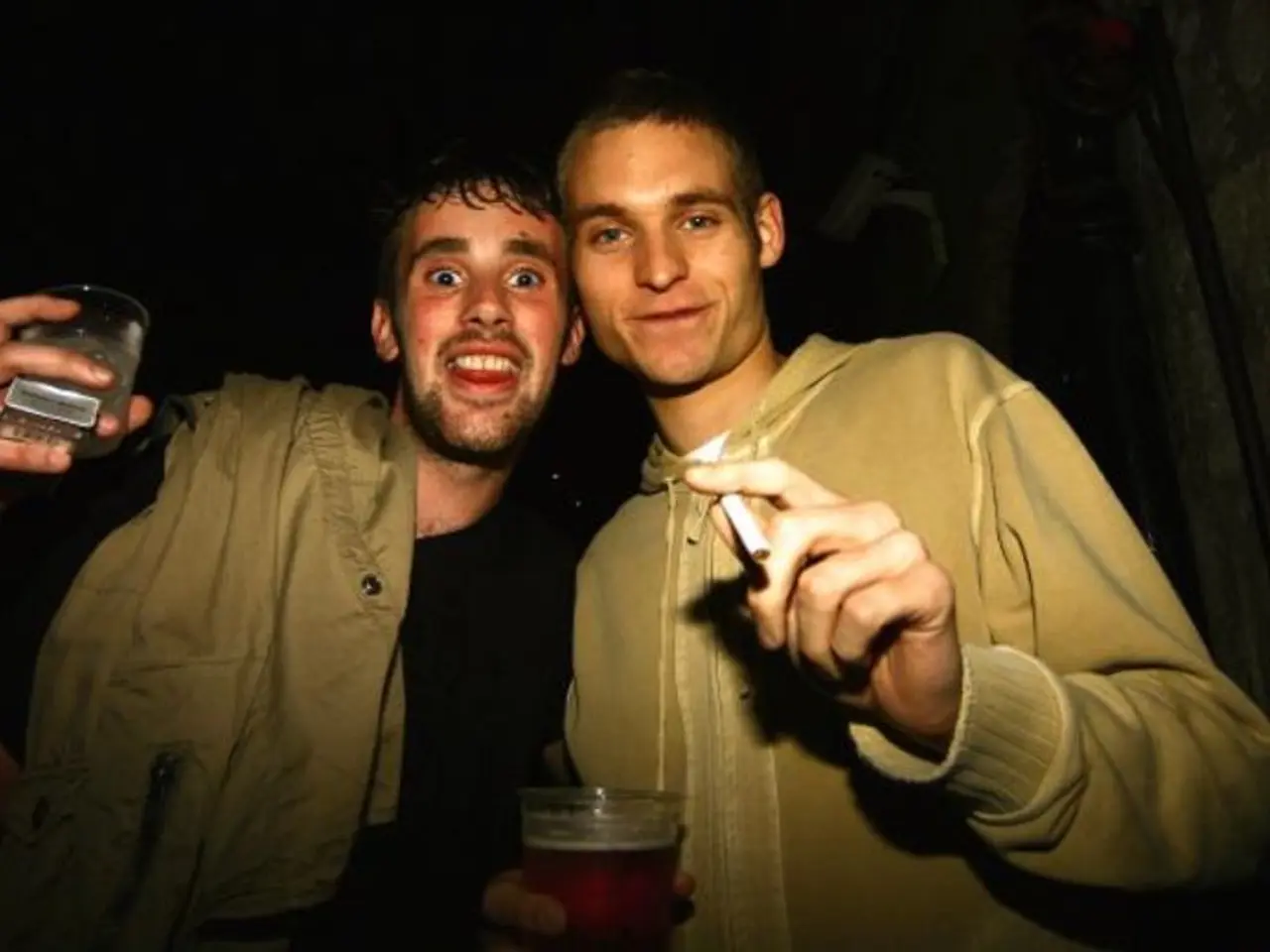 There are two men standing holding glass in one hand and cigarette in another hand.