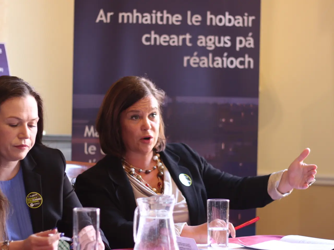 In this image I can see two women wearing black colored blazers are sitting on chairs in front of a...