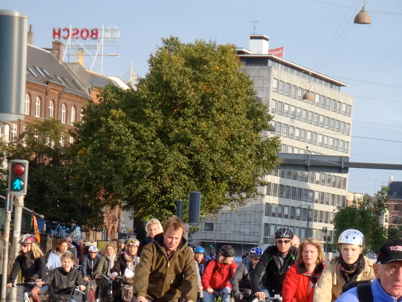 In this image there are people cycling on a road, in the background there are poles, trees,...