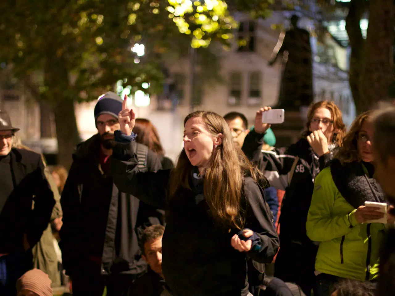 This image consists of many persons. In the middle, we can see a woman shouting. She is wearing a...