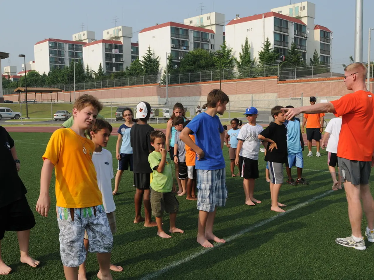 Land is covered with grass. Here we can see people. Background there are buildings, light poles,...
