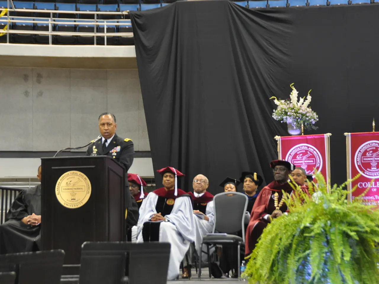 In this picture we can see a plant in the front, there are some people sitting on chairs and a man...