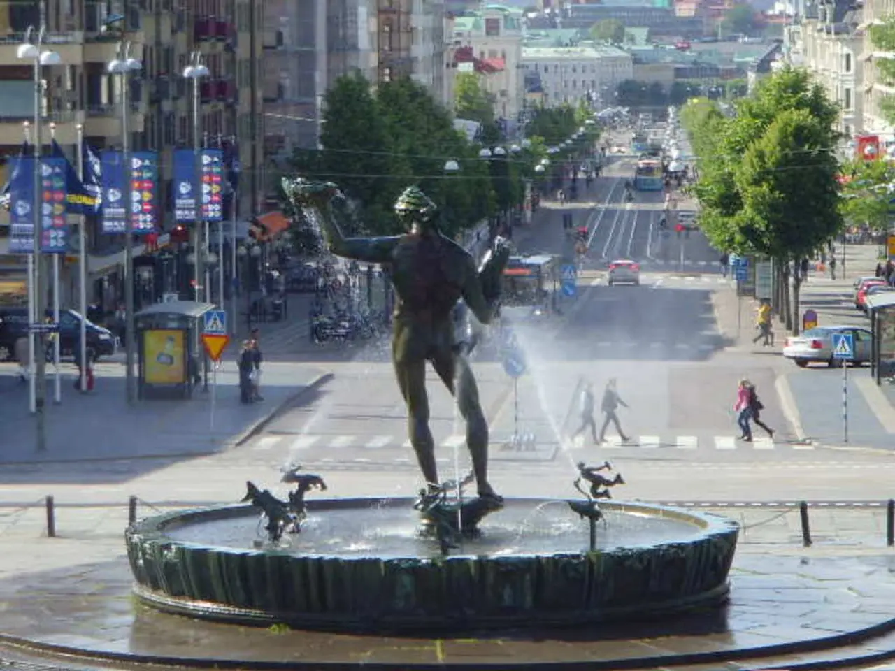 In this image we can see a fountain, there are some buildings, trees, boards, poles, lights, people...