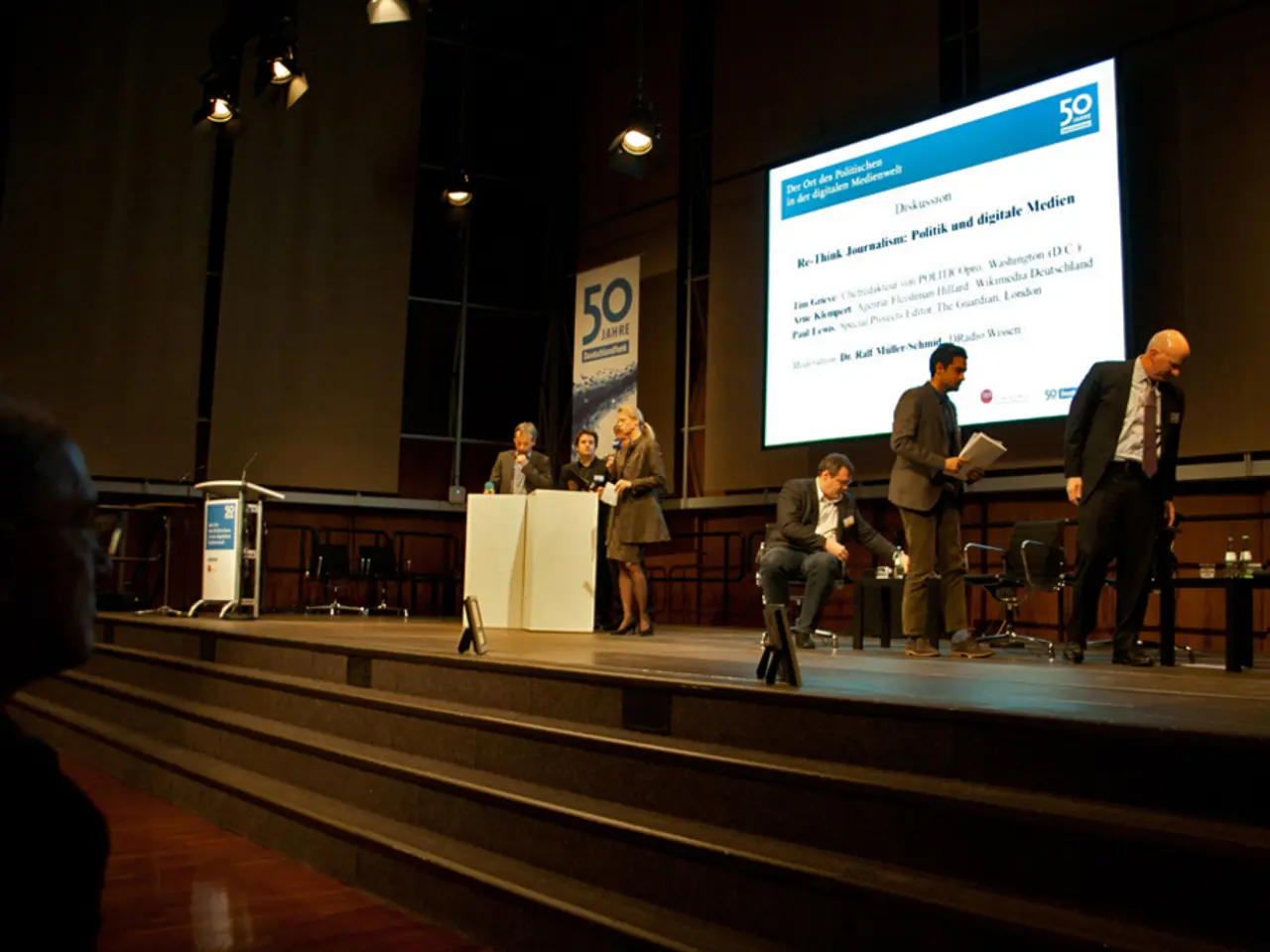 There is a stage with steps. On the stage there are chairs and tables. There are many people...