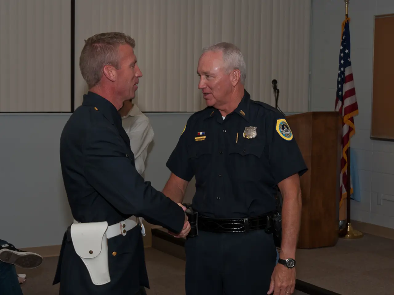 In this picture I can see 2 men in front and I see they're shaking hands and I see they're wearing...