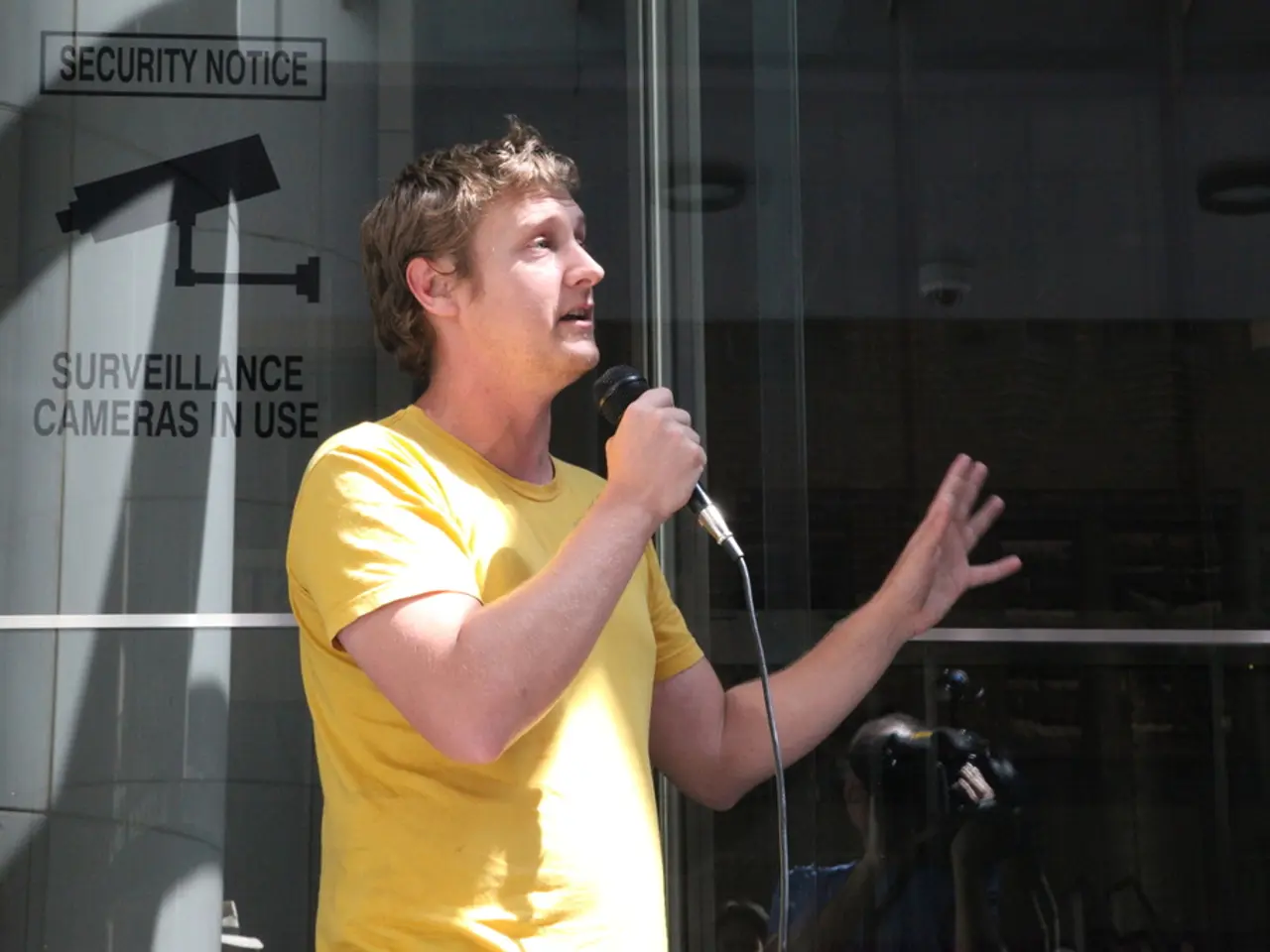 A person is talking on mic. Behind him there is a glass door.