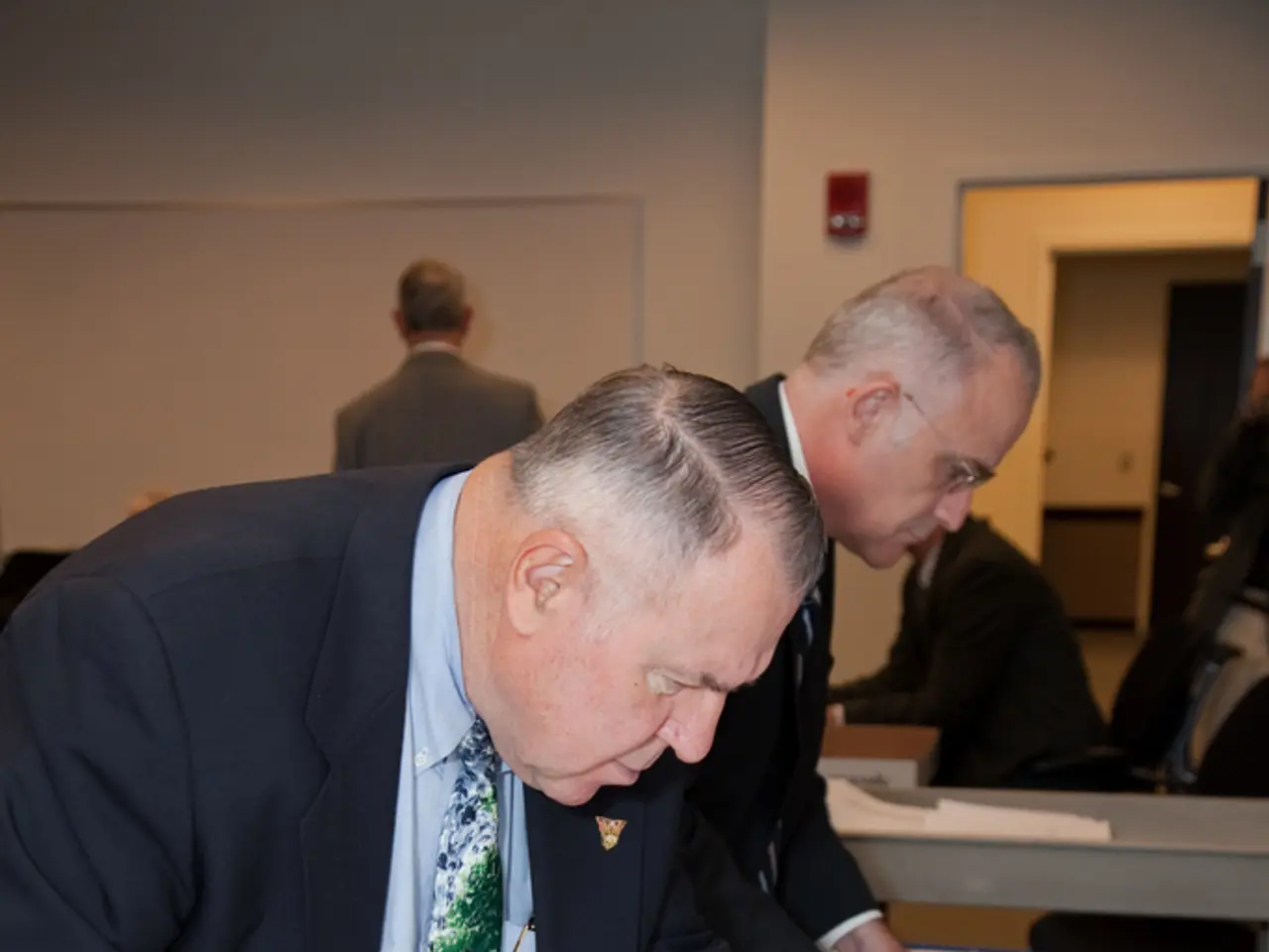 In this image I see 2 men who are wearing suits and I see that this man is holding papers and I see...