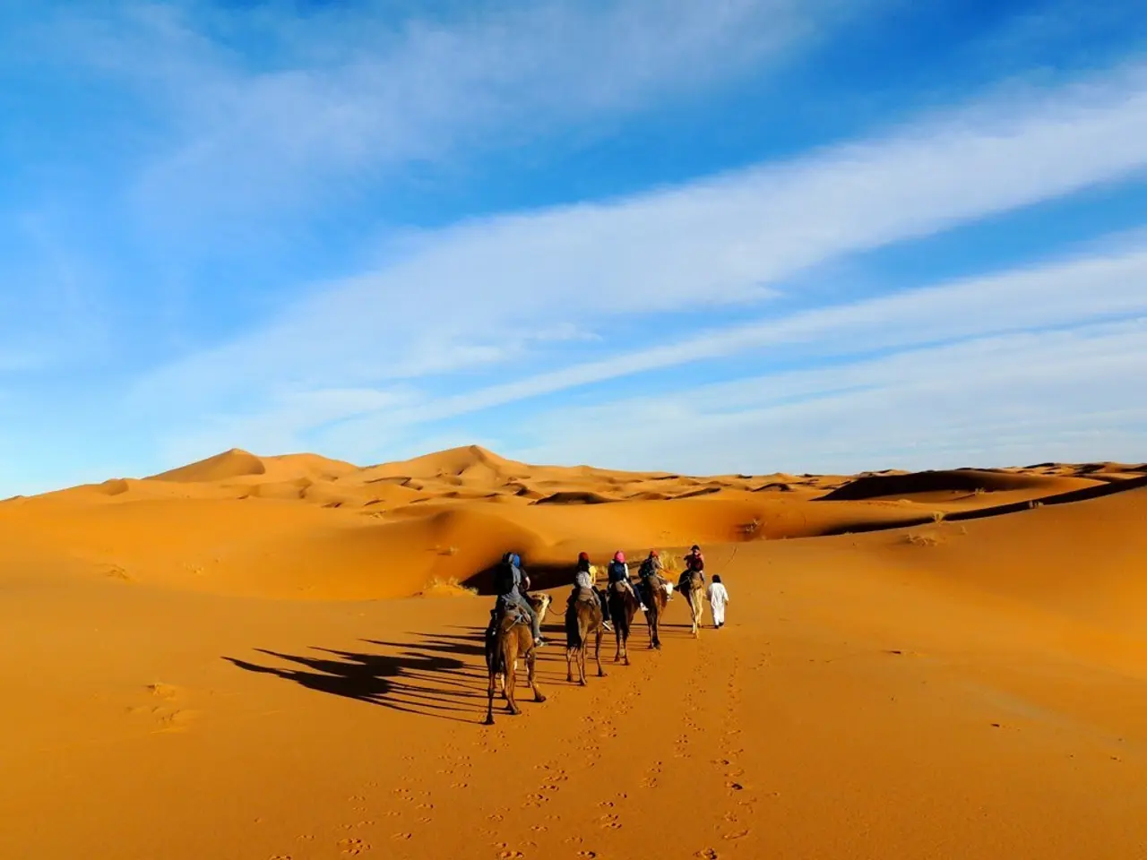 In this image there is a desert, five people riding camels on the desert and a person is walking,...
