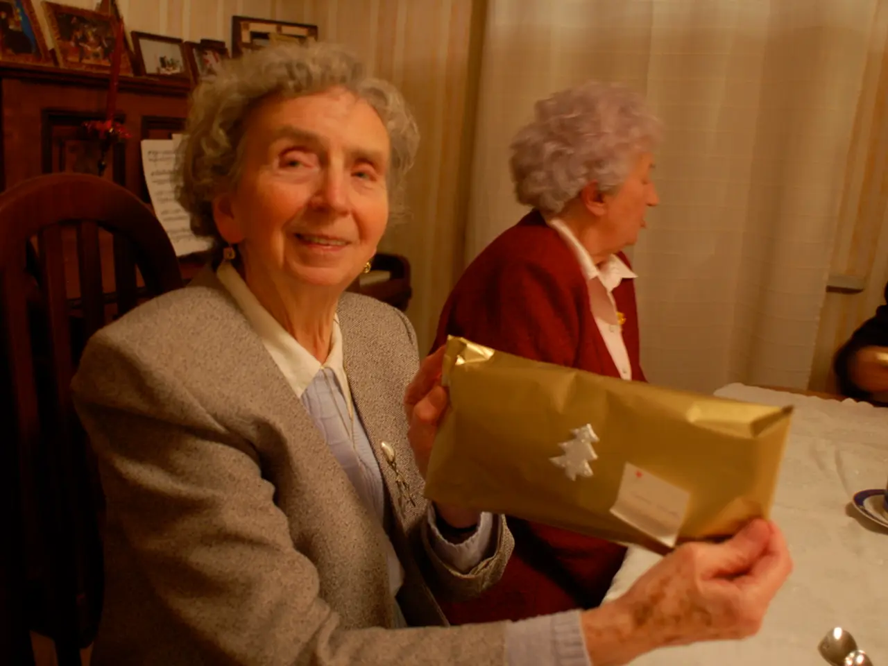 In this picture there is a woman sitting, smiling and showing a gift pack and another woman beside...