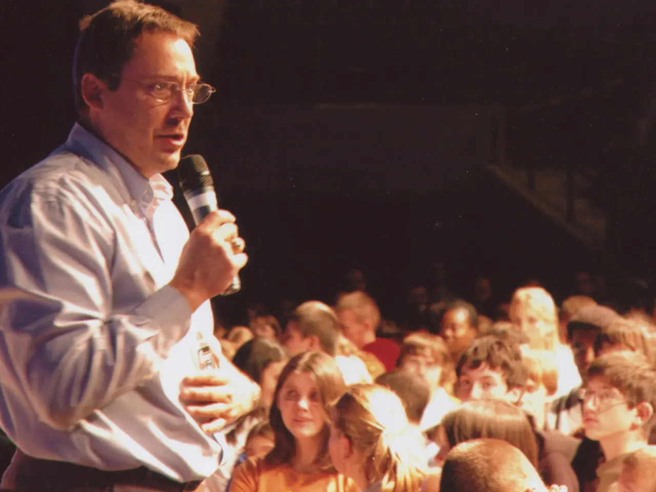 In this picture there is a man wearing a blue color shirt is giving a speech. Beside there is a...