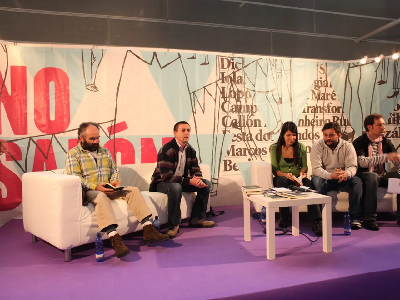 In the image there are few men and women sat on sofa on a stage,in front there is a table on which...