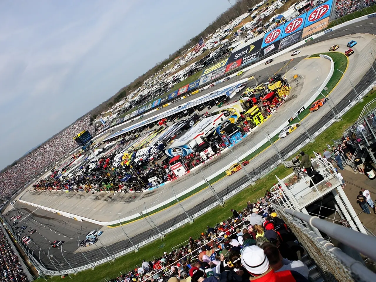In this image, we can see a crowd. There is a racing track and some cars in the middle of the...