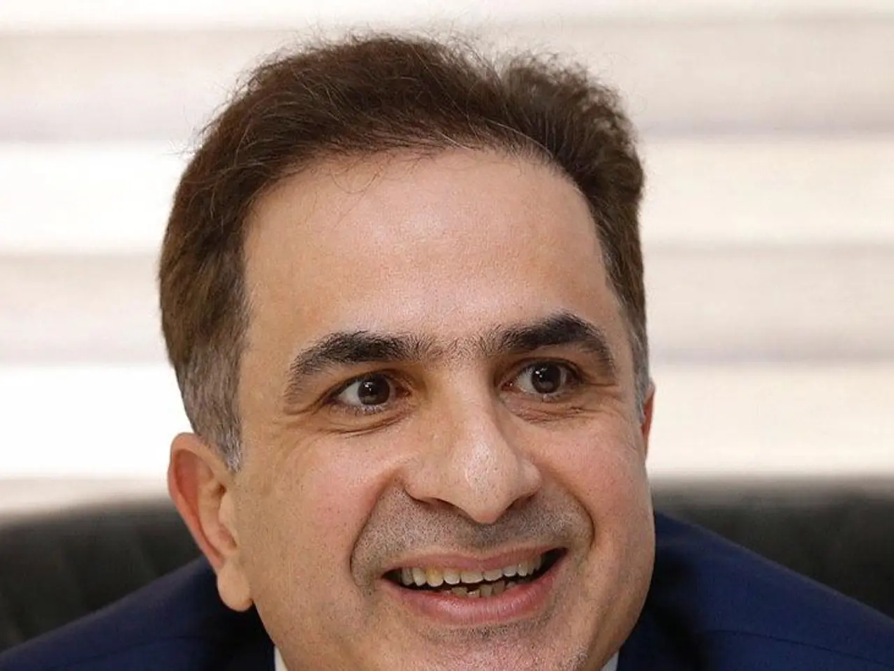 The image shows a man in a blue suit smiling at the camera against a blurred background. He appears...