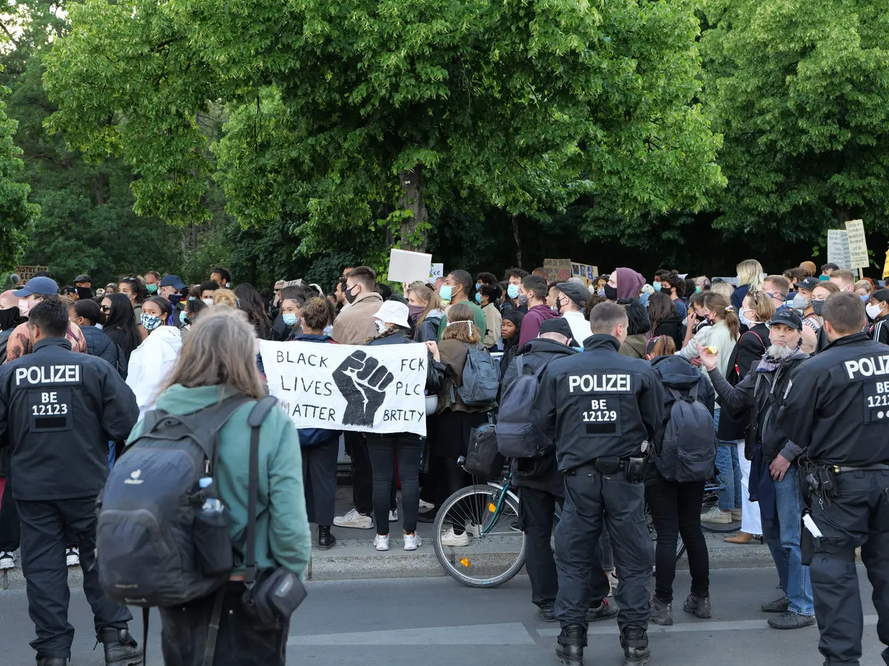 The image shows a large group of people standing on the side of a road, some of them holding...