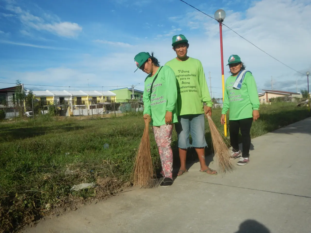 The image shows three people wearing green t-shirts and caps, sweeping the street with brooms. On...
