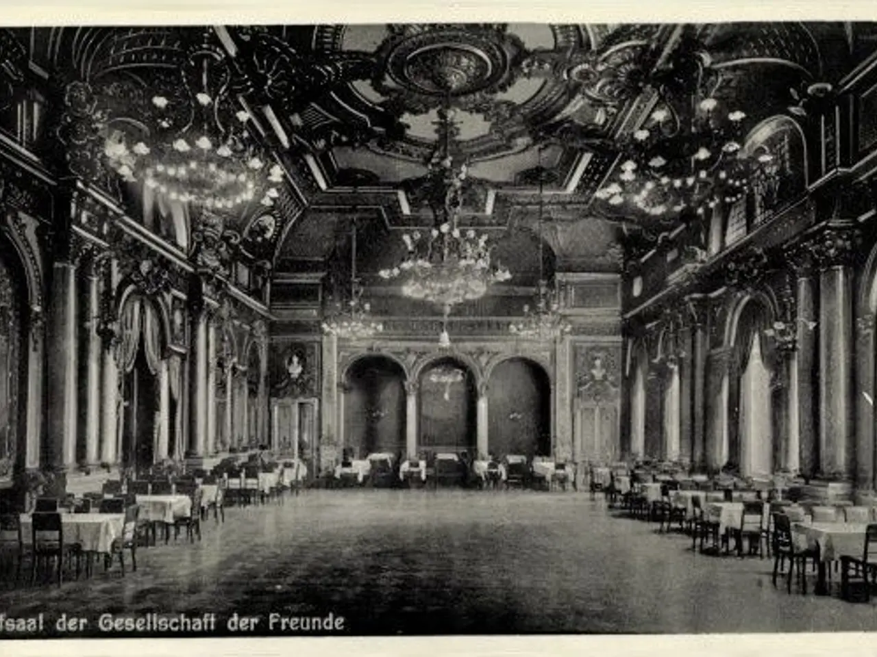 The image shows an old black and white photo of a large room with tables and chairs, pillars,...