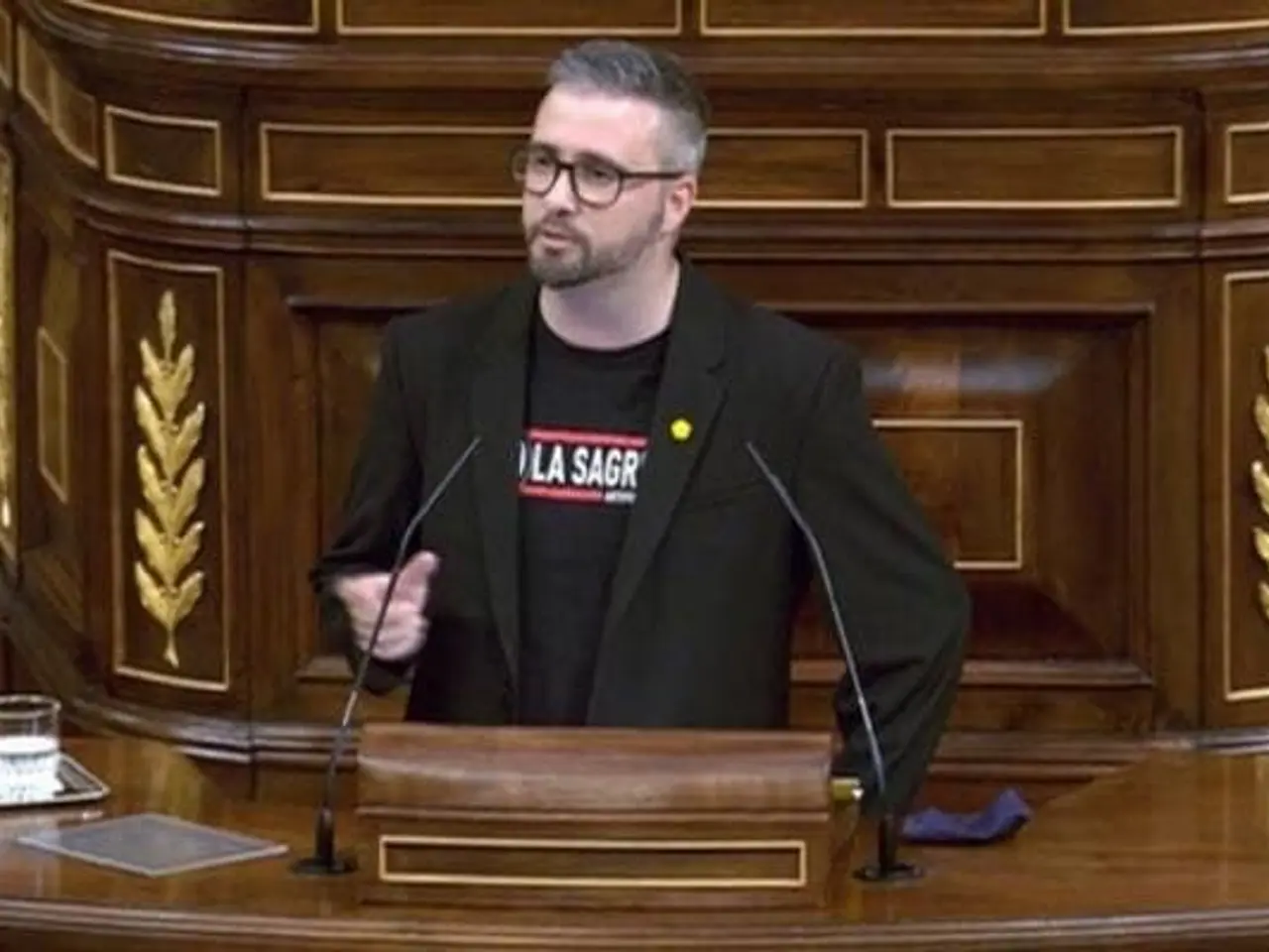 The image shows a man standing at a podium in front of a wooden wall, wearing a black blazer and...