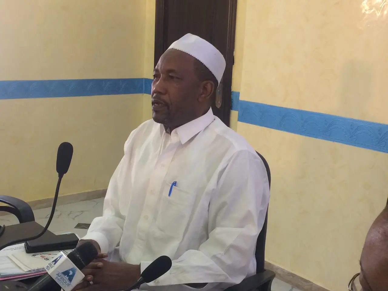 The image shows a man sitting at a table with microphones in front of him, wearing a white dress...