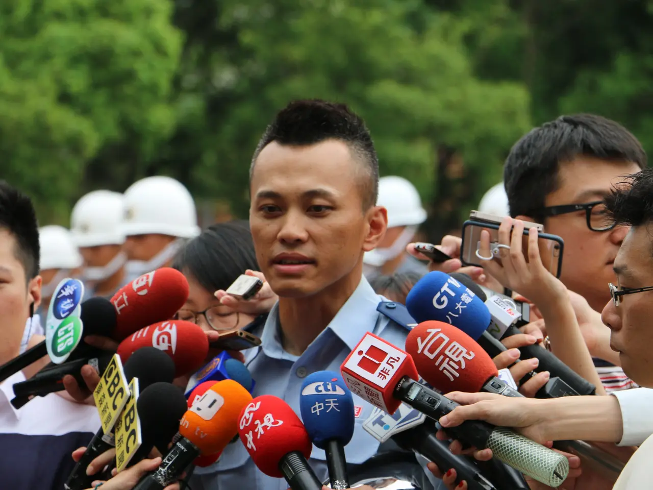 The image shows a man in a police uniform surrounded by reporters, some of whom are holding mics...