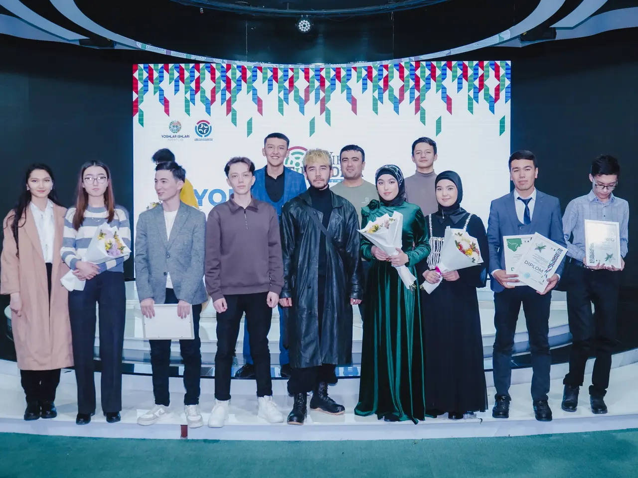 The image shows a group of people standing on top of a stage, each holding a bouquet of flowers and...