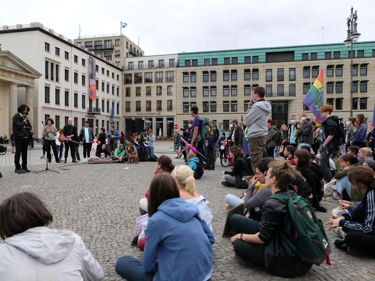 The image shows a group of people sitting on the ground in front of a crowd of people, some of whom...