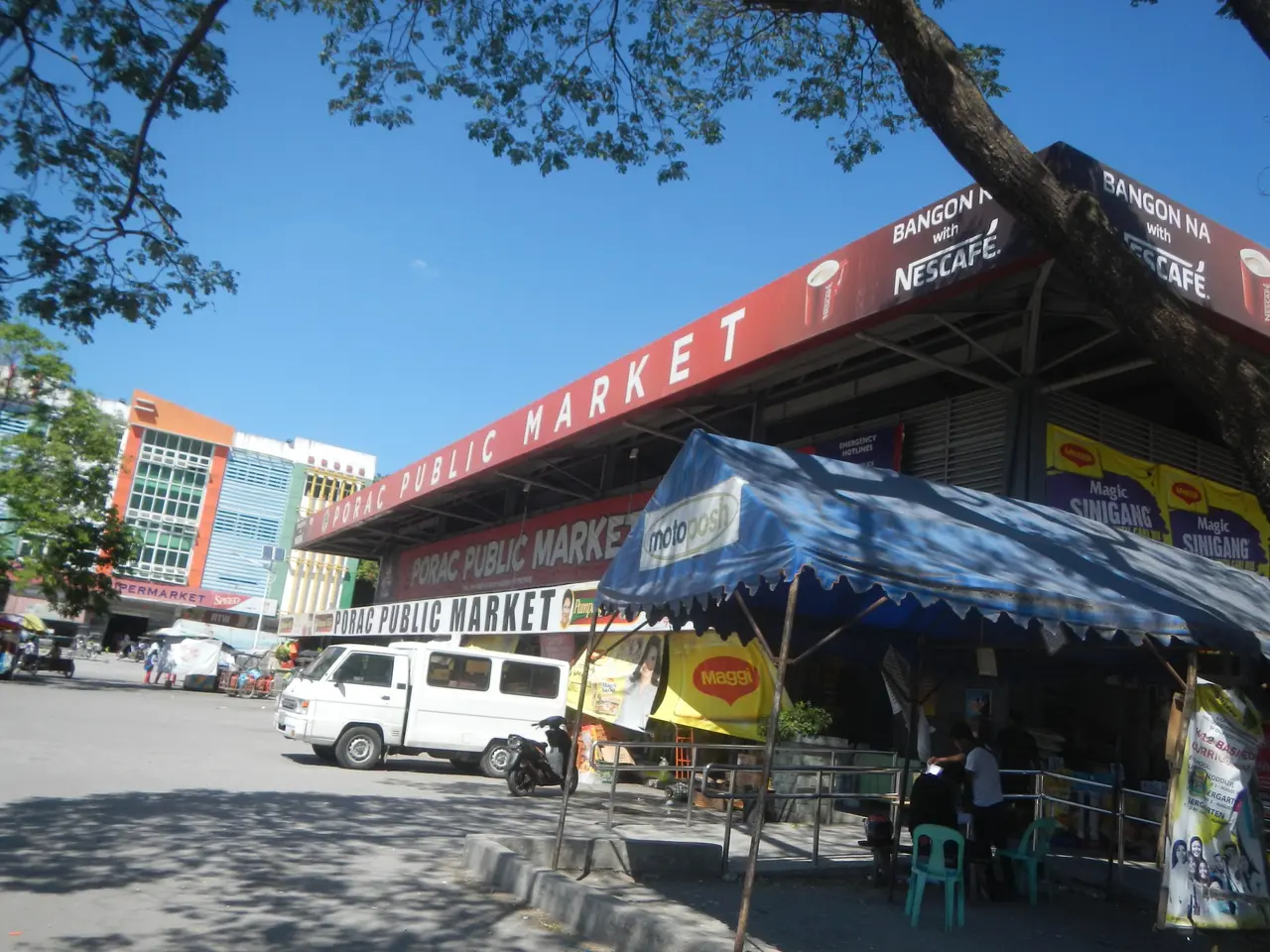 The image shows a bustling public market in the middle of a city street, with buildings, trees,...