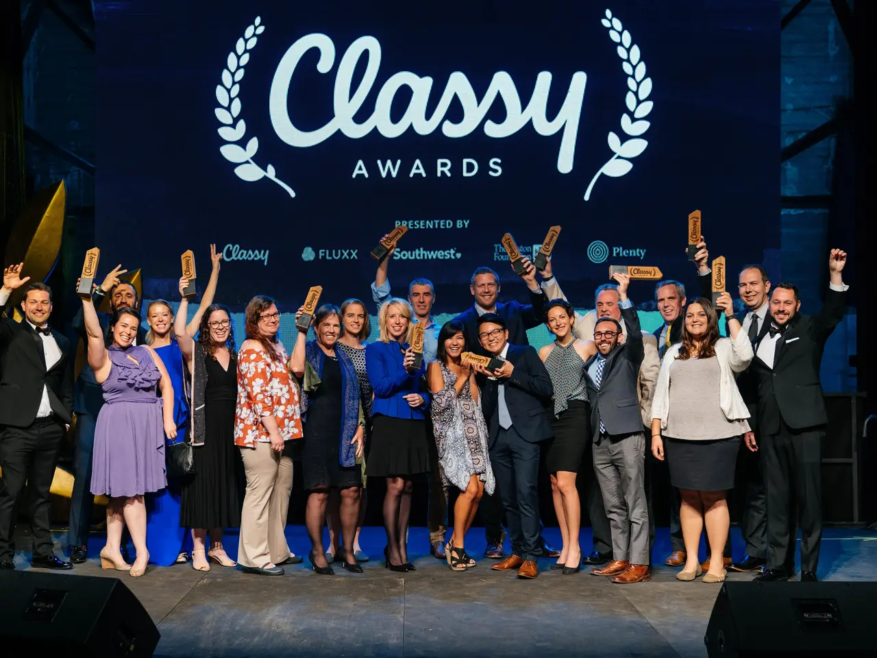 The image shows a group of people standing on top of a stage, smiling and holding awards in their...