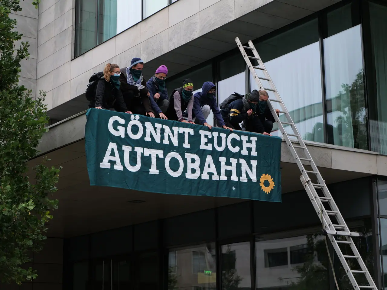 The image shows a group of people standing on top of a building, holding a banner with text on it....