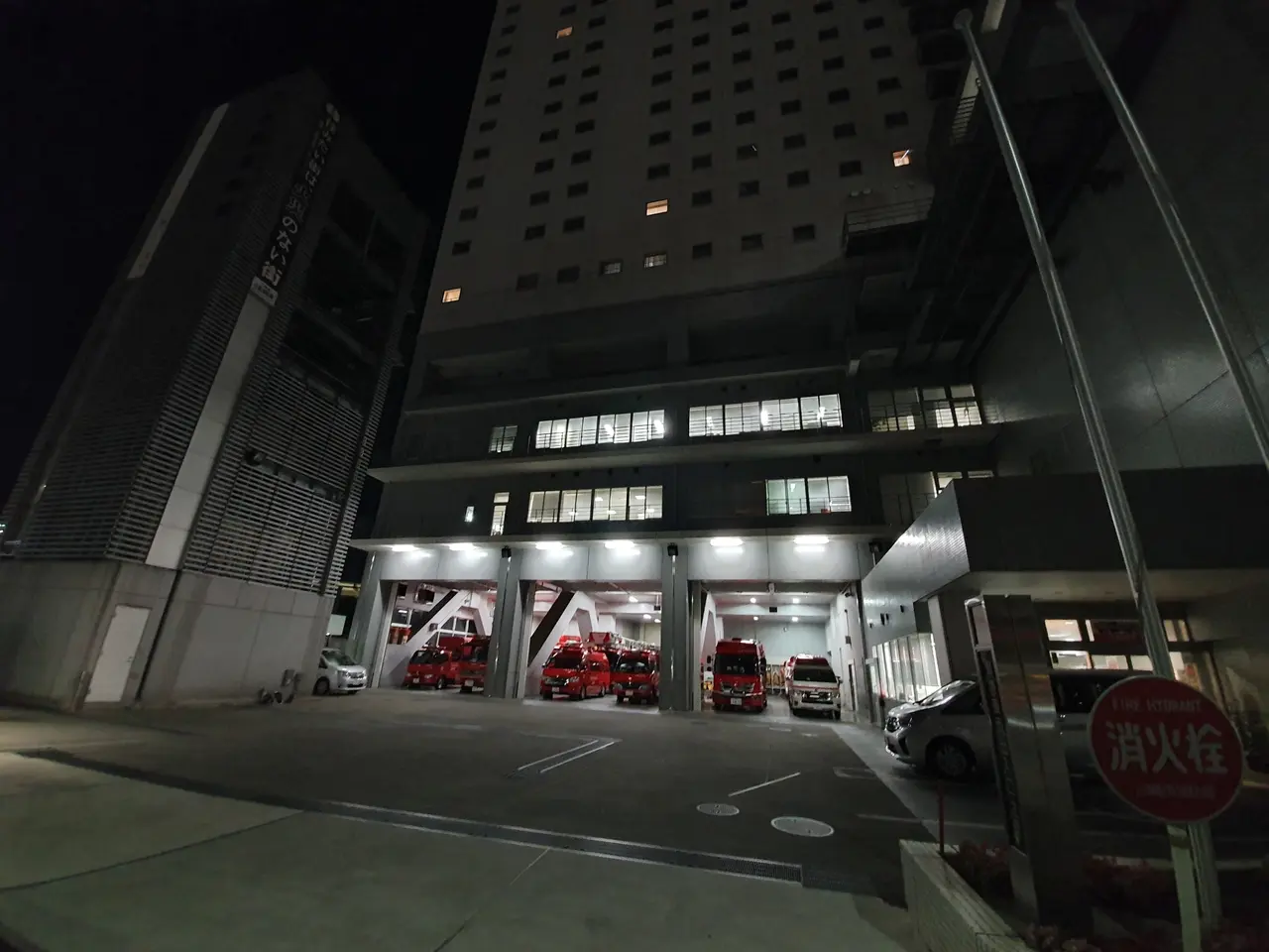 The image shows a fire station at night with several fire trucks parked in front of it. The...