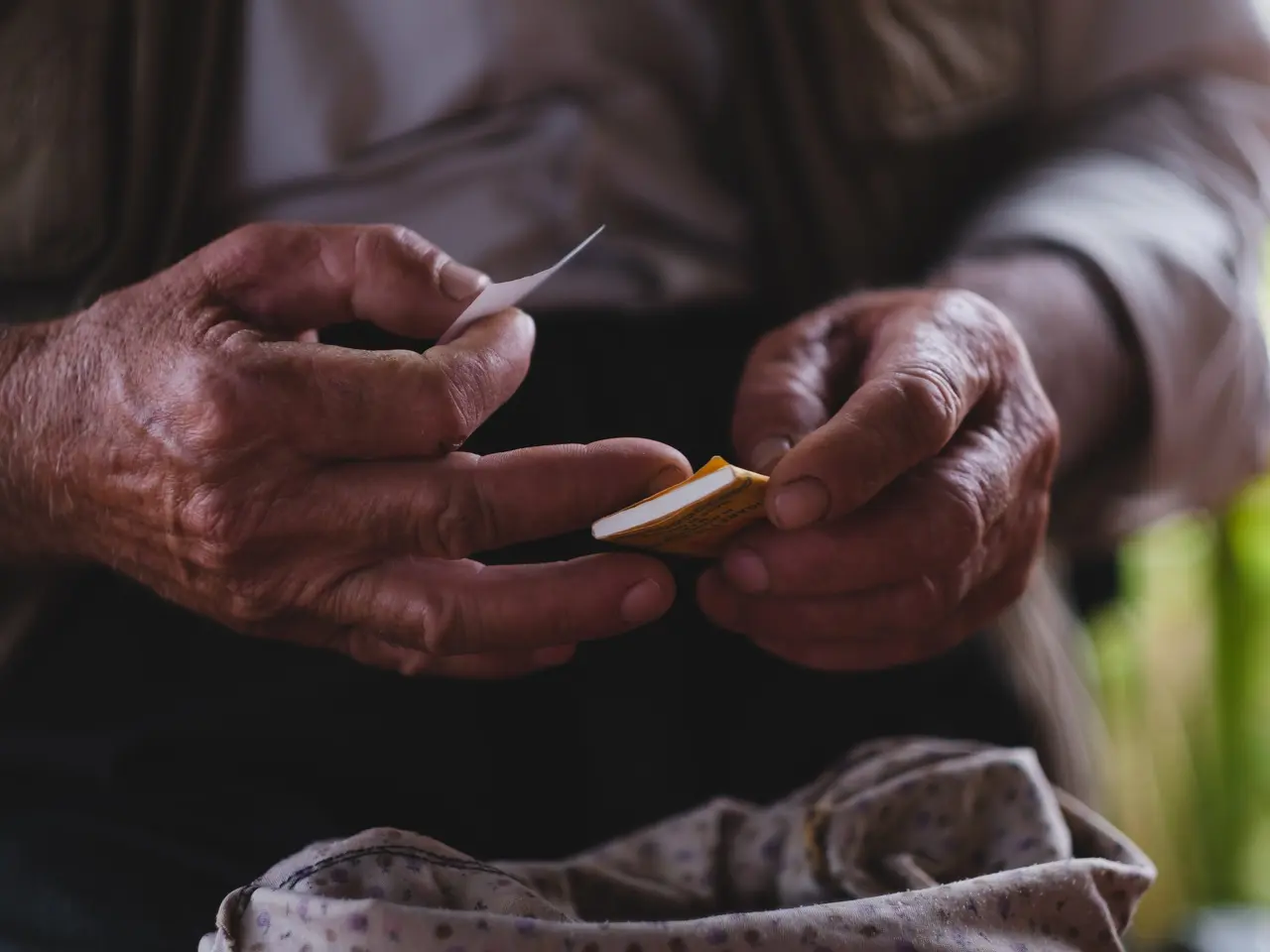 The image shows an elderly man holding a piece of paper in his hands, with a blurred background. He...