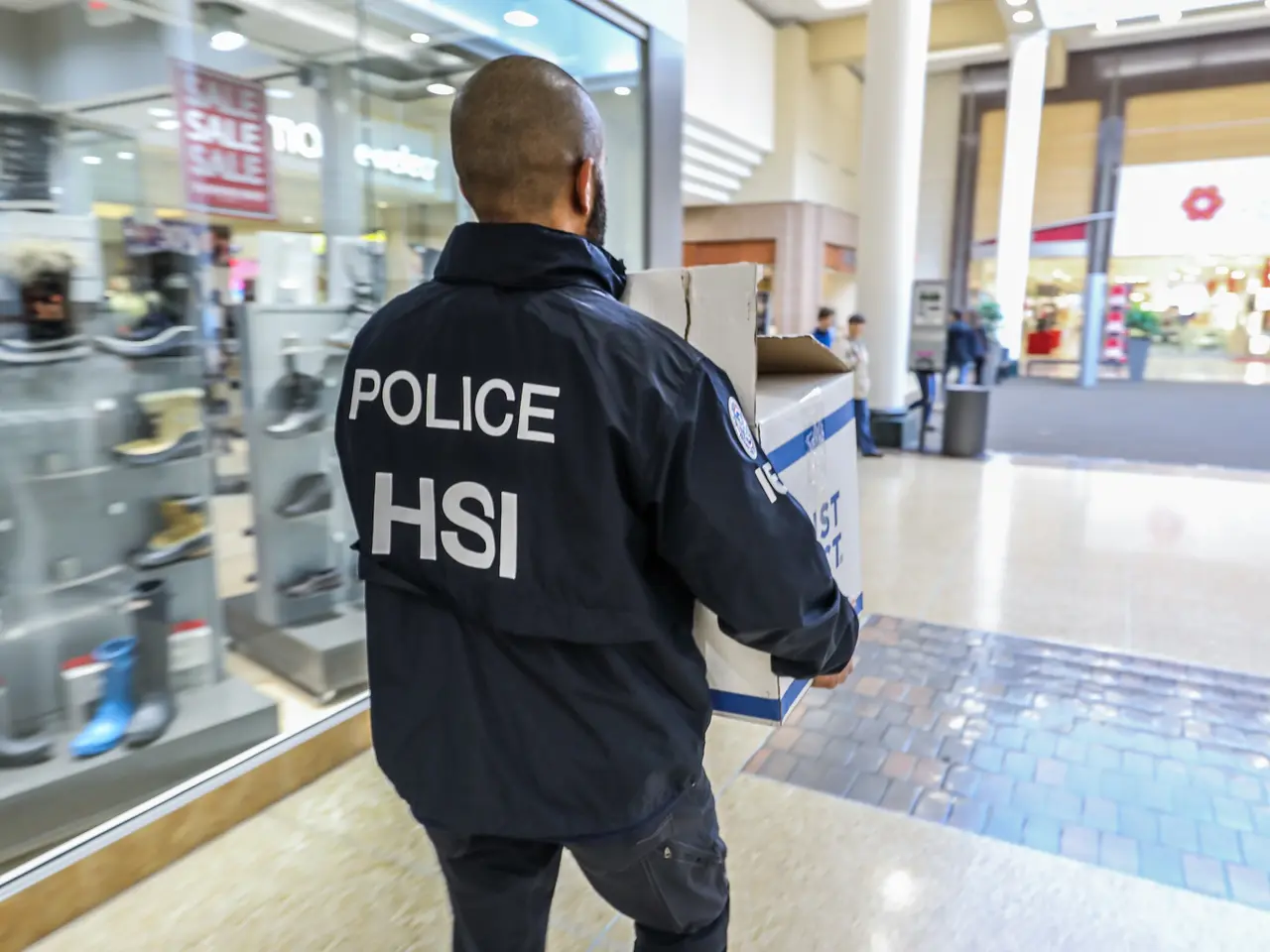 The image shows a police officer walking through a shopping mall, carrying a box in his hand. He is...