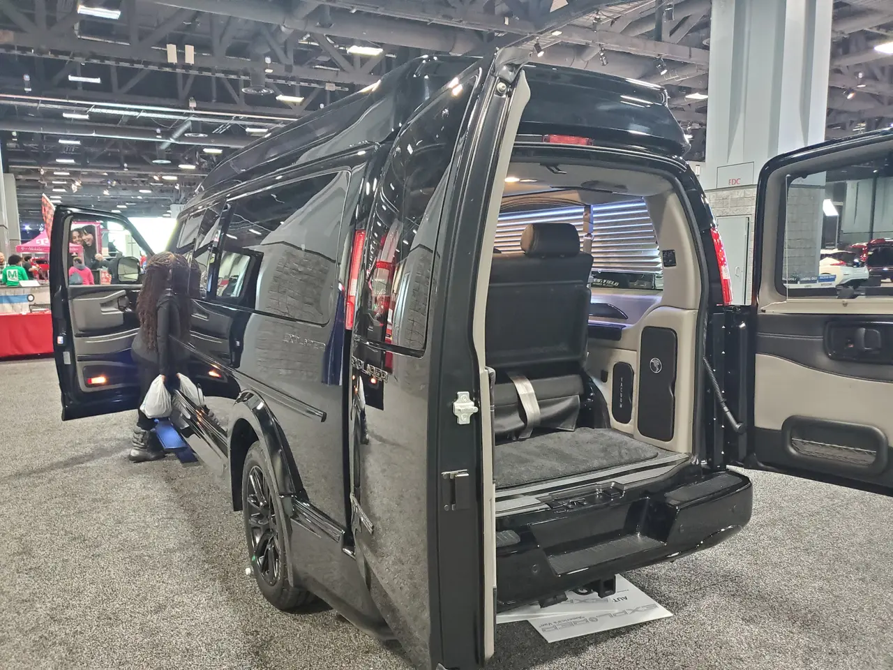 The image shows a black van with its doors open on display at a show, surrounded by a group of...