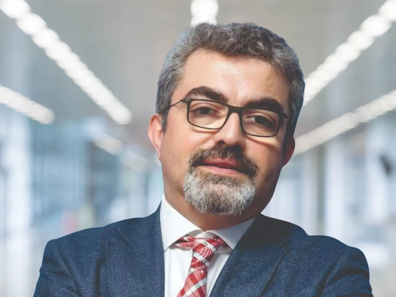 The image shows a man in a suit and tie with his arms crossed, wearing spectacles and standing in...