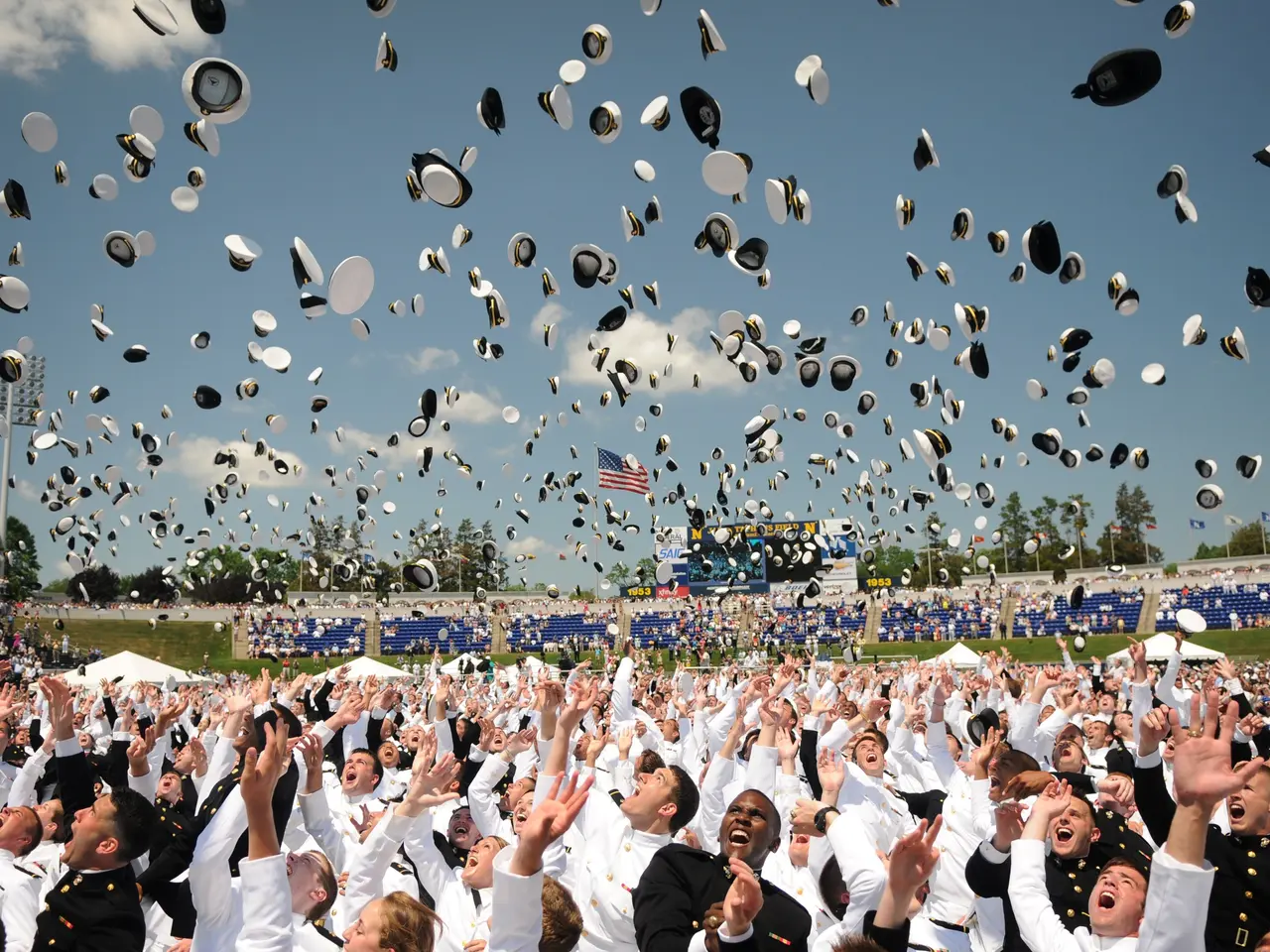 The image shows a large group of people in white graduation caps and gowns throwing their hats in...