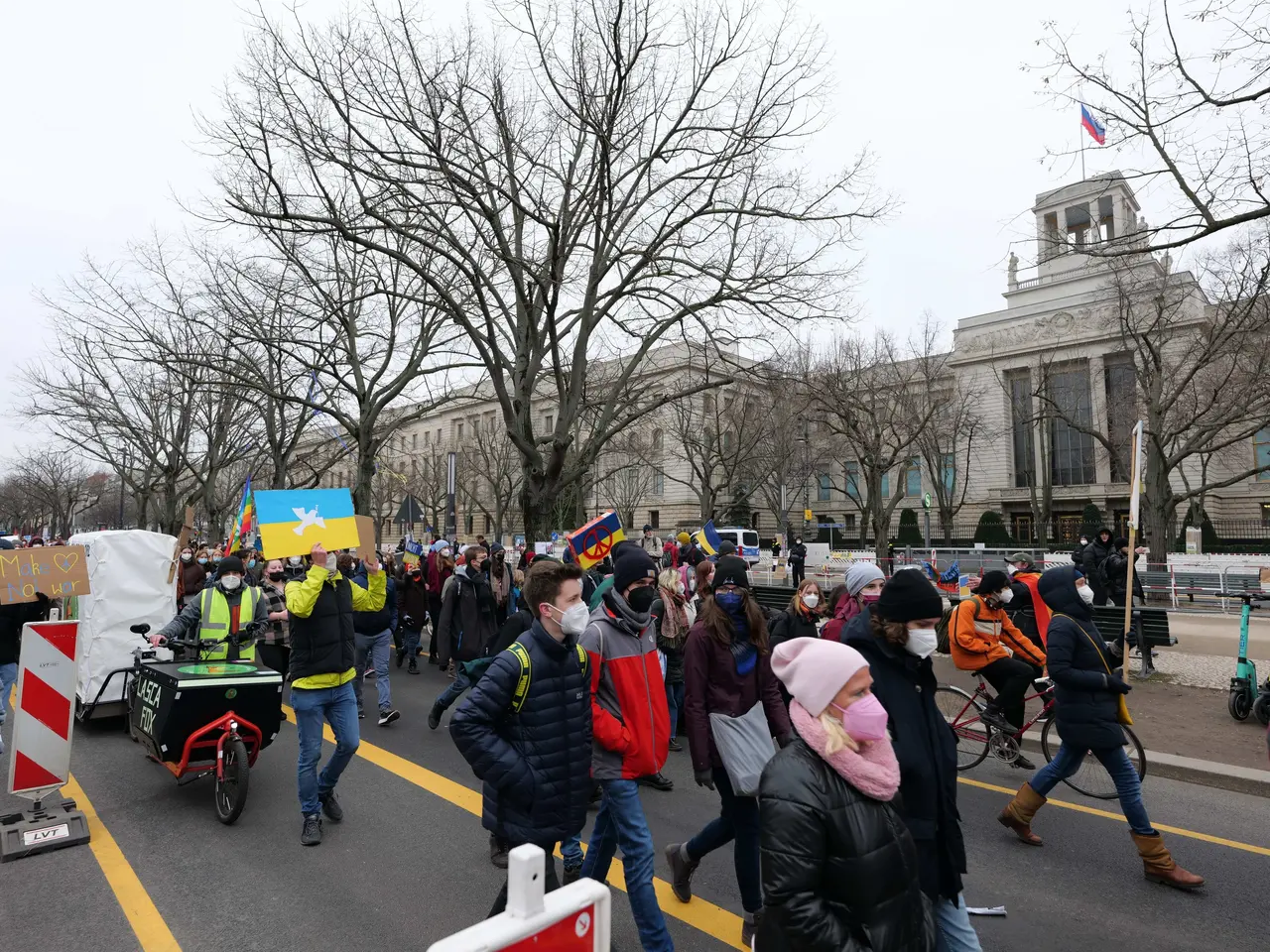 The image shows a large group of people walking down a street in front of a building, some of them...