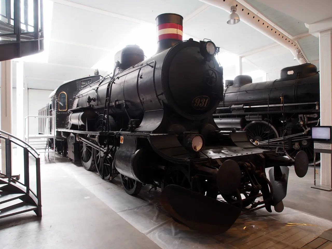The image shows an old steam locomotive on display in a museum, surrounded by a floor, railings,...